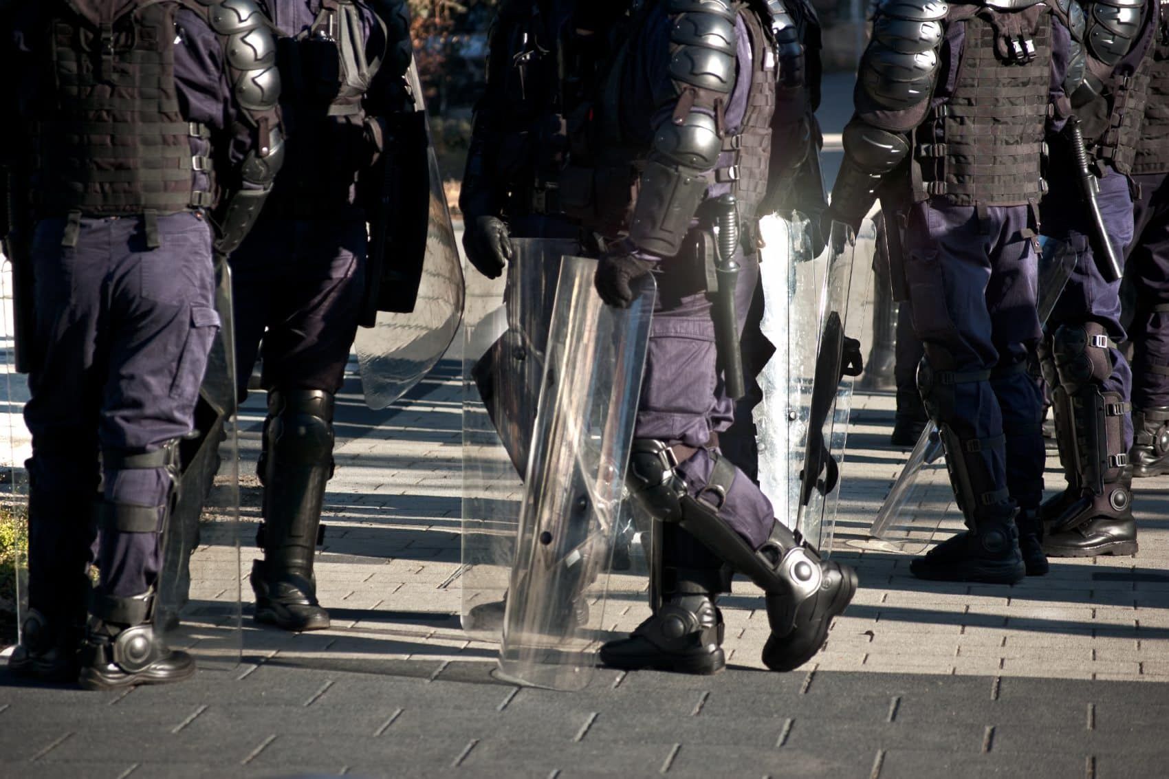 A group of officers with shields and riot gear are shown from the shoulders down - police brutality