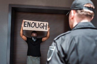 Black protester with mask on and holding "Enough" sign faces white officer - police brutality