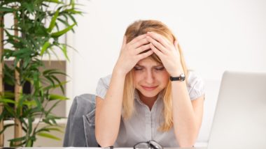 A sad woman sits at a desk with her head in her hands, with a laptop, glasses and paperwork in front of her - Facebook moderator