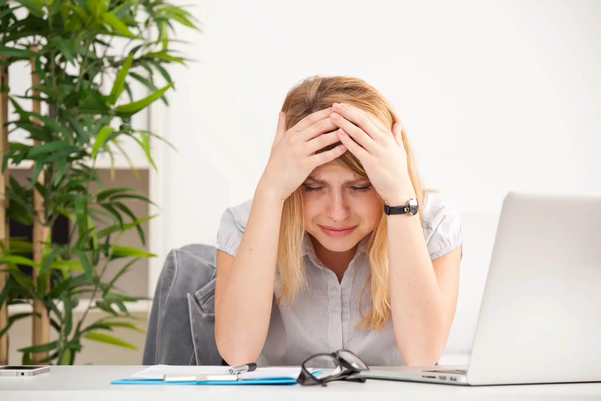 A sad woman sits at a desk with her head in her hands, with a laptop, glasses and paperwork in front of her - Facebook moderator