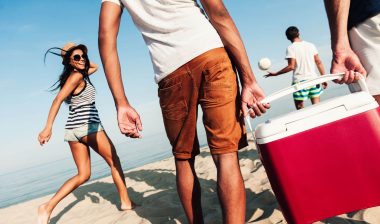 Two men walking with a group of beachgoers carry a red cooler with a white lid - Igloo coolers
