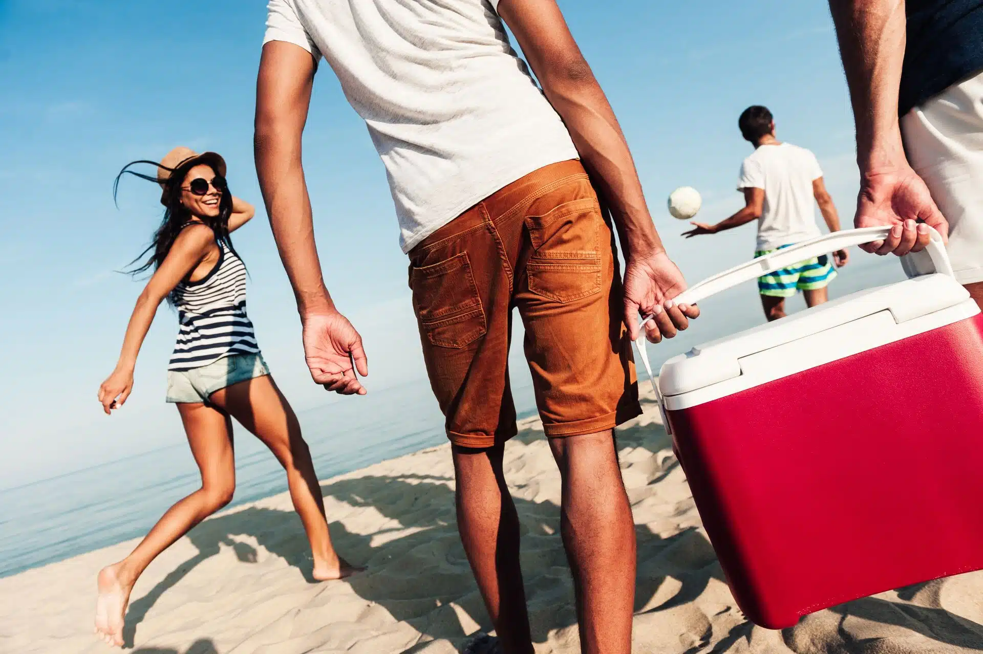 Two men walking with a group of beachgoers carry a red cooler with a white lid - Igloo coolers