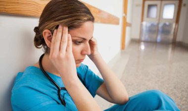 A nurse in blue scrubs sits on a hospital hallway floor, rubbing her temples - coronavirus outbreak