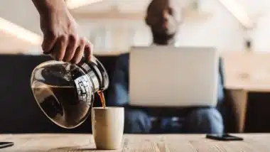 Someone pours coffee for man sitting near a table while working on a laptop - Maxwell House