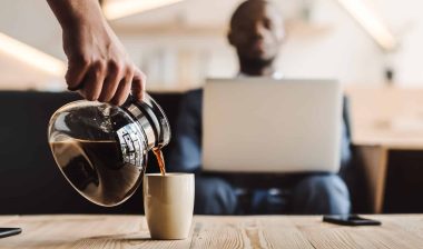 Someone pours coffee for man sitting near a table while working on a laptop - Maxwell House