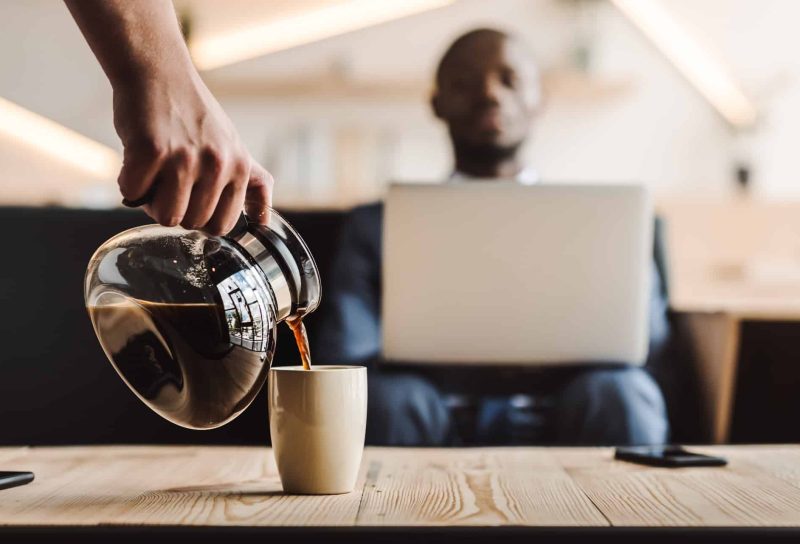 Someone pours coffee for man sitting near a table while working on a laptop - Maxwell House