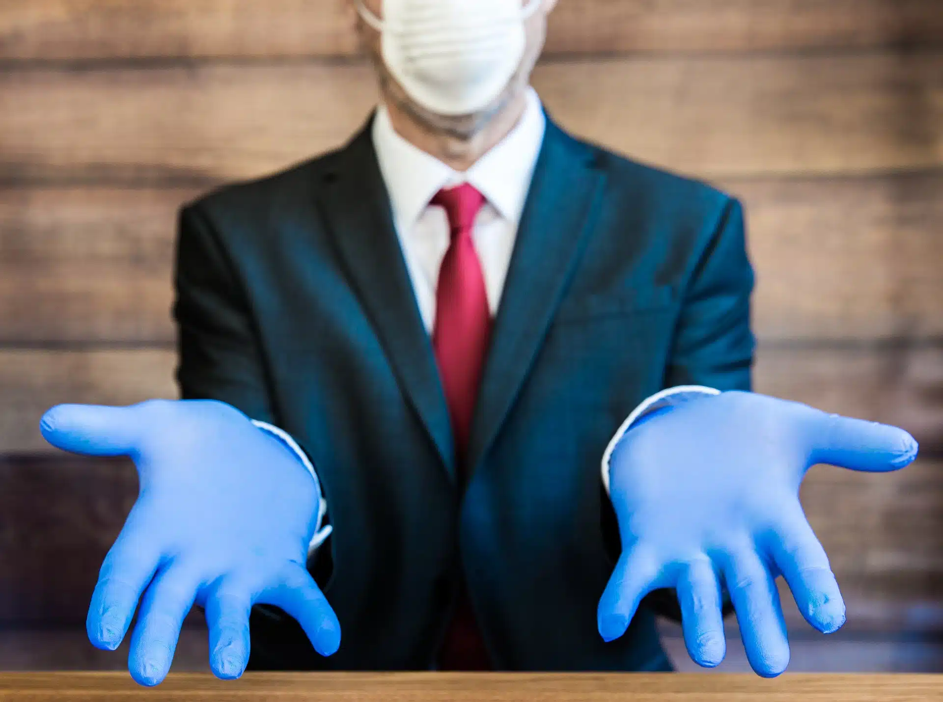 A man in a suit sits at a table and holds out his hands, wearing blue gloves and a white face mask - ppe shortage