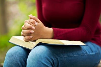 A woman prays outdoors with her hands folded on top of a book in her lap - Capitol Hill Baptist Church