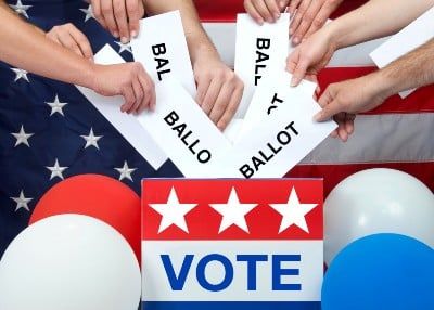 Hands reach to put ballots in a box marked "vote" near balloons and in front of an American flag - arkansas voters