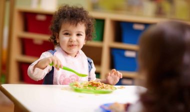 A happy young child in a bib eats while sitting at a classroom table - Feeding Our Future
