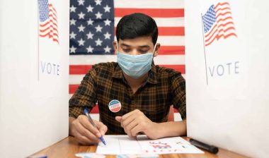 A man sits in a polling booth to vote while wearing a mask, with an American flag backdrop - arkansas voters