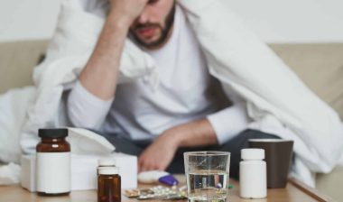 An ill man sits with his head in his hand, with medicine bottles and a glass of water on the table in front of him - coronavirus cure