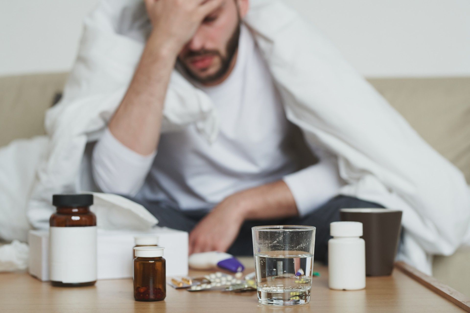 An ill man sits with his head in his hand, with medicine bottles and a glass of water on the table in front of him - coronavirus cure