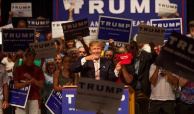 Donald Trump holds up a red "Make America Great Again" hat at a rally - Trump campaign