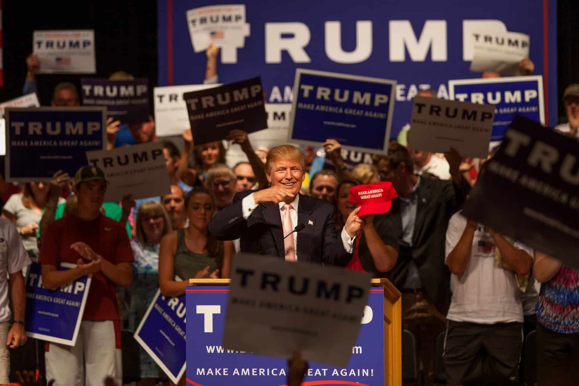 Donald Trump holds up a red "Make America Great Again" hat at a rally - Trump campaign