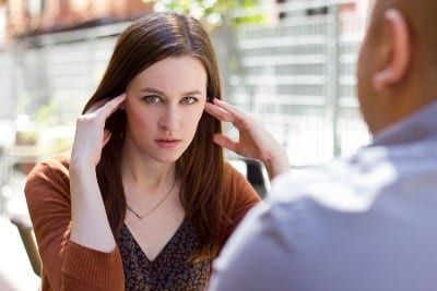 A woman on a bad date looks at the camera while holding her temples - It's Just Lunch