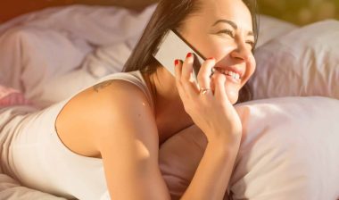 A smiling woman talks on an iPhone while lying on her stomach in bed - iPhone radiation