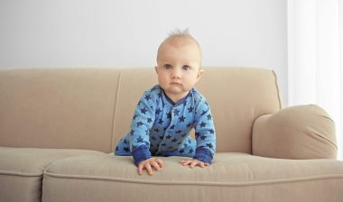 A baby wearing a blue romper crawls on a tan sofa - target recalls