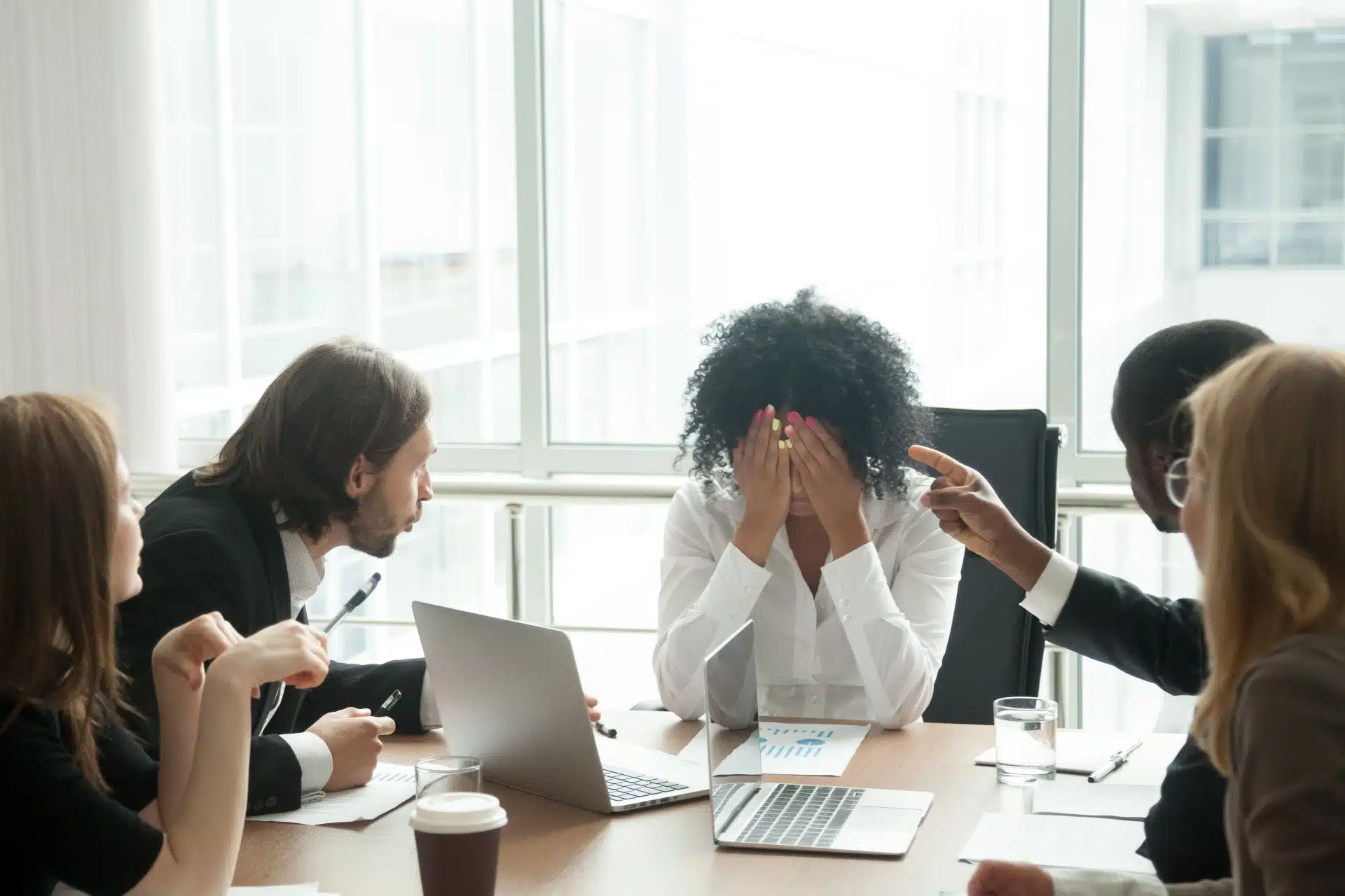 A Black woman at the end of a conference table holds her head in her hands as employees talk at her - racial discrimination