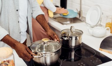 Man handles stainless steel pot on stove as woman washes dishes in the background - All-Clad stainless steel
