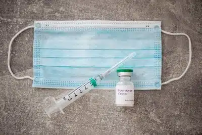 A syringe and vial of coronavirus vaccine lie on top of a blue surgical mask 