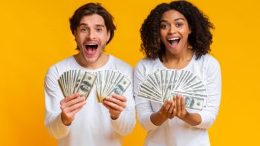 A man and woman hold fanned-out cash at arm's length in front of them as they stand against a yellow background