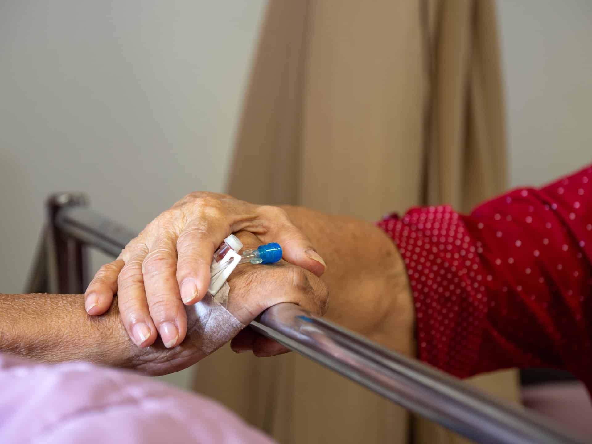 An elderly woman holds the hand of an elderly man lying in a hosptial bed - See's Candies worker