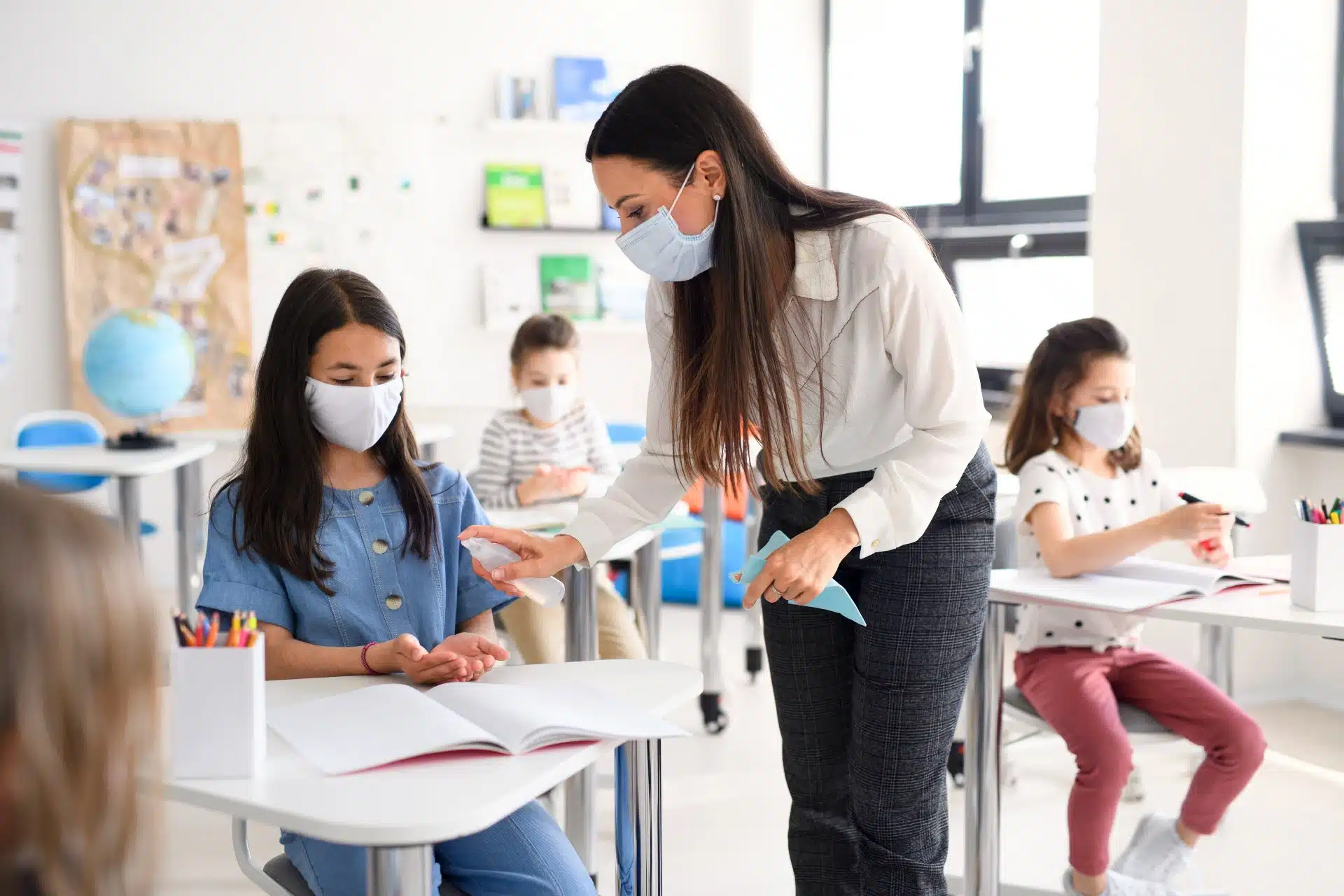 A teacher in a face mask distributes some hand sanitizer to a student in a classroom