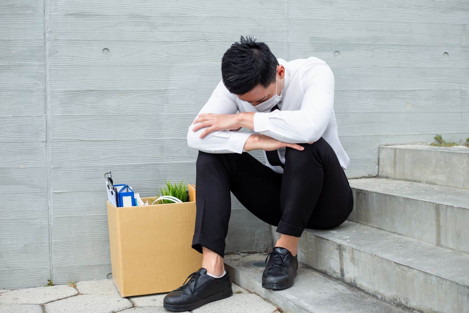 A man wearing black and white sits near a box of office contents on some concrete steps with his head hanging - mass layoffs