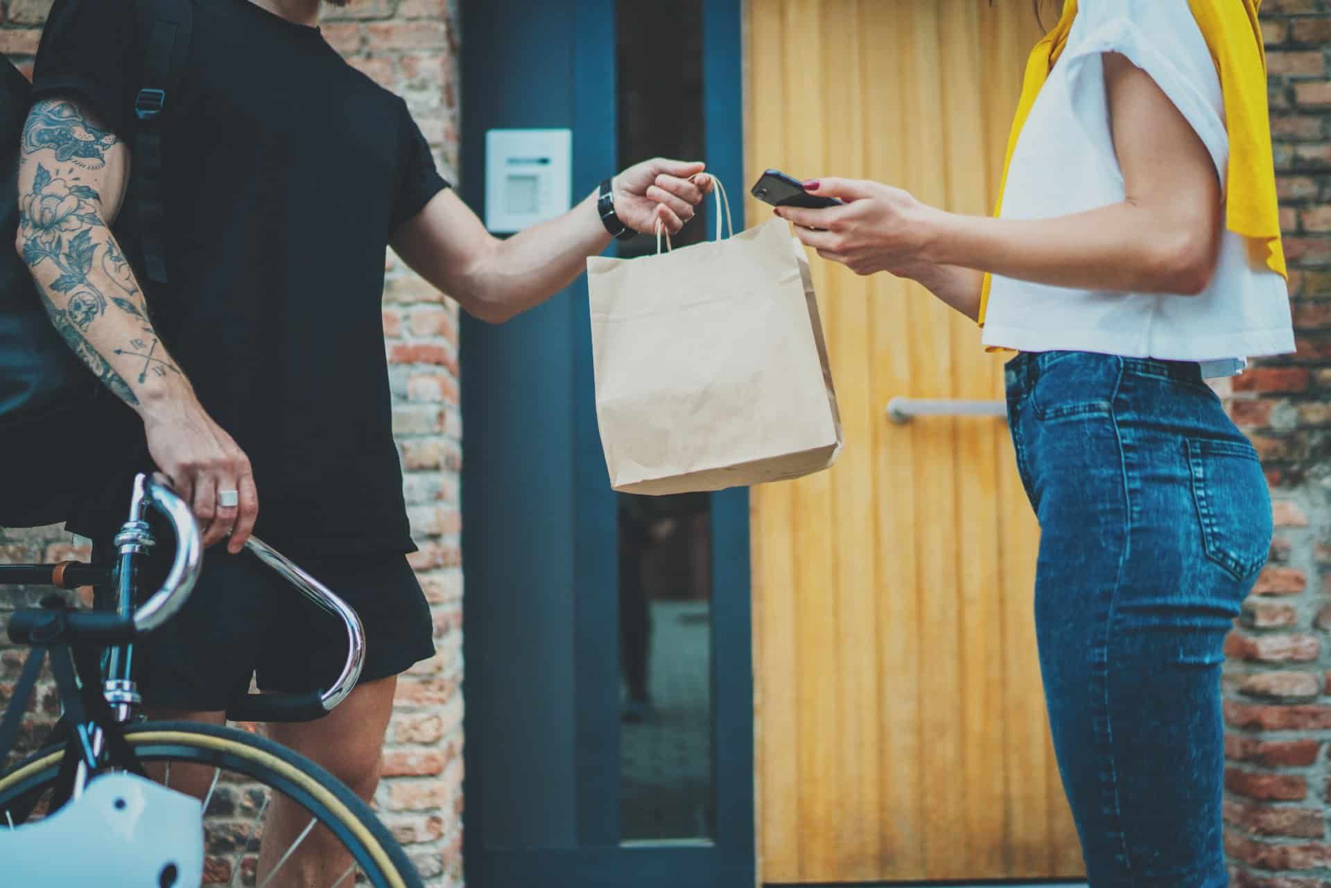 A tattooed courier delivers food in a brown bag to a woman with a smartphone - food delivery