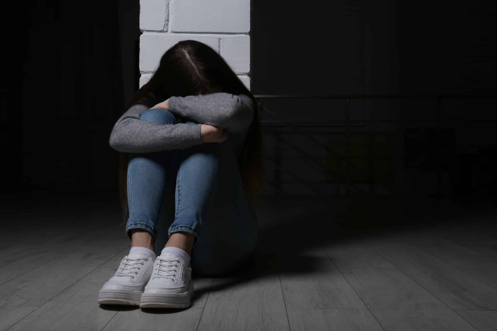 Teen girl hides face behind arms as she sits on the floor near a pillar with her knees pulled to her chest - Boys & Girls Club of America
