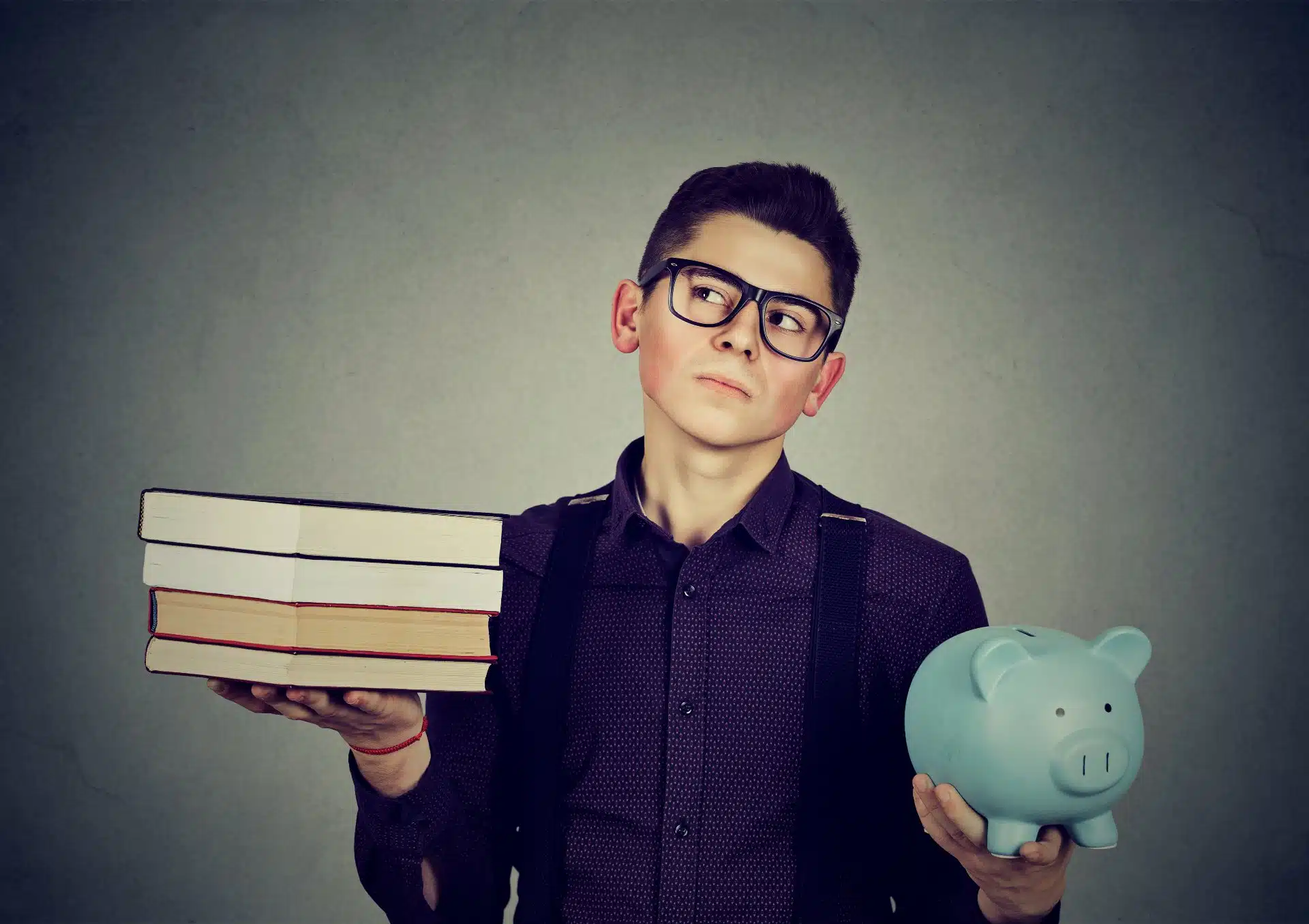 A young man wearing glasses and a backpack holds a stack of four books in his right hand and a blue piggy bank in his left hand - pheaa