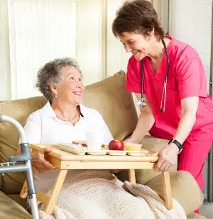 A health care worker in pink scrubs brings a meal tray to an elderly woman — humana