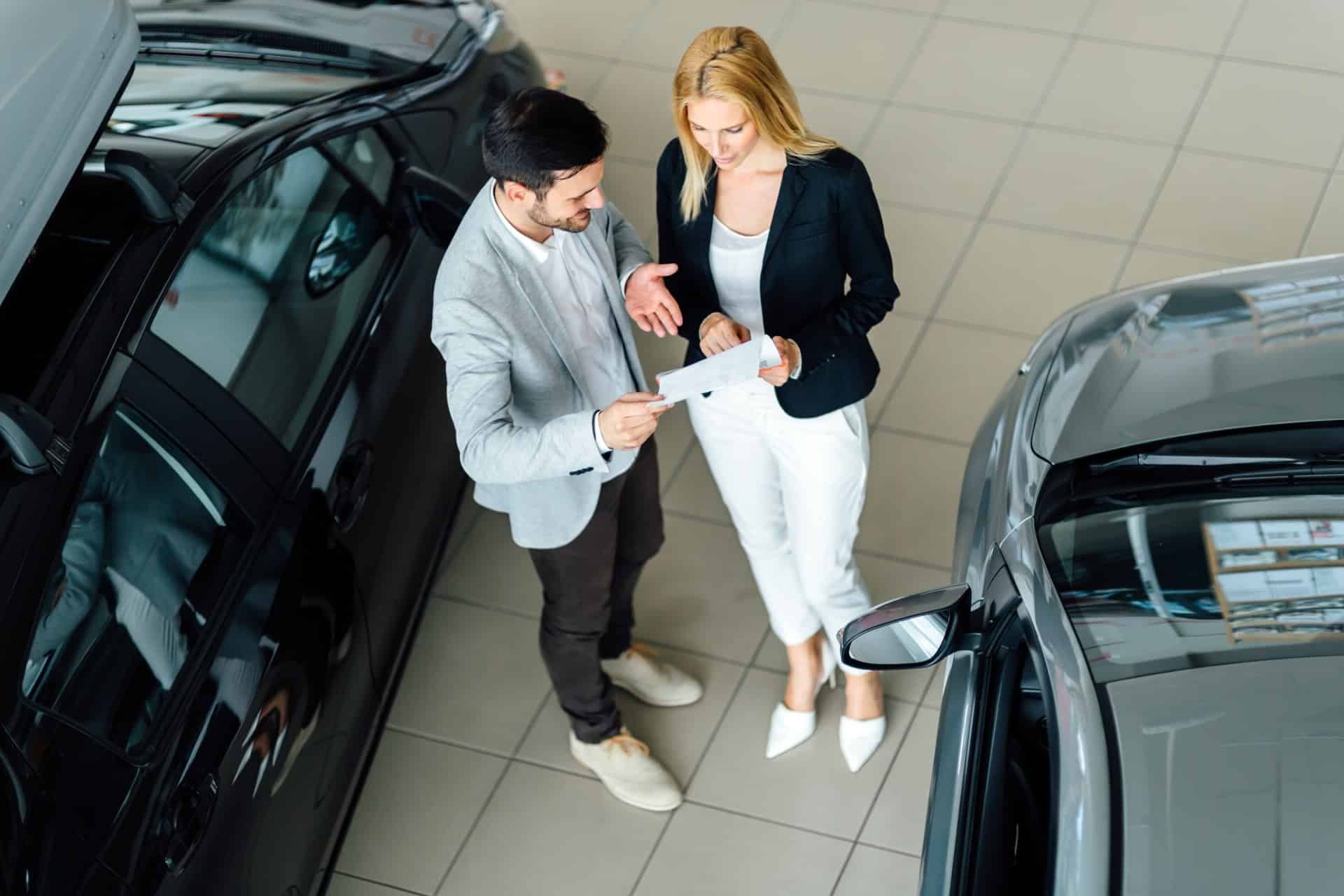 A salesperson and a customer talk over a paper while standing in a car dealership showroom - reynolds