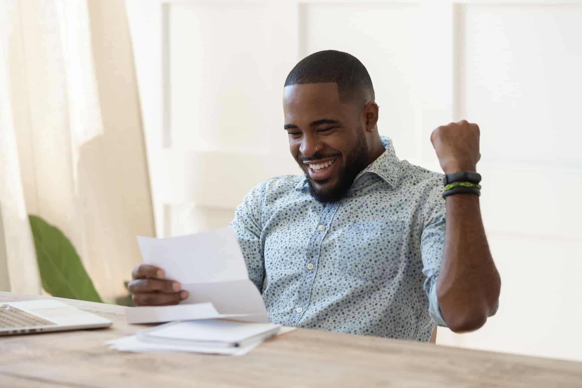 A happy man pumps his fist as he reads a letter while sitting at a desk.