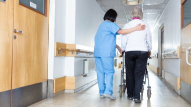 A nurse in blue scrubs helps a patient walk down a hallway at a nursing facility or hospital - Prompt Nursing