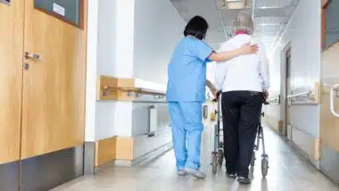 A nurse in blue scrubs helps a patient walk down a hallway at a nursing facility or hospital - Prompt Nursing