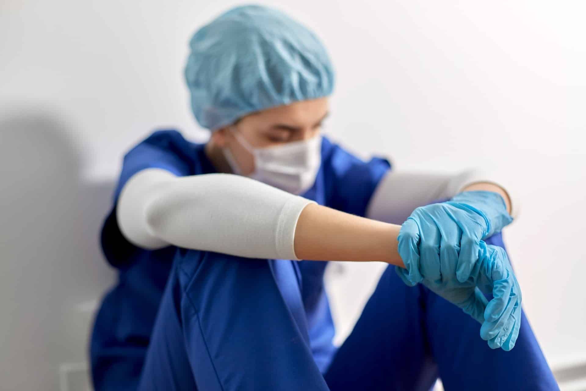 A nurse in blue scrubs and protective gear sits on the floor with her arms on her knees - chi franciscan health