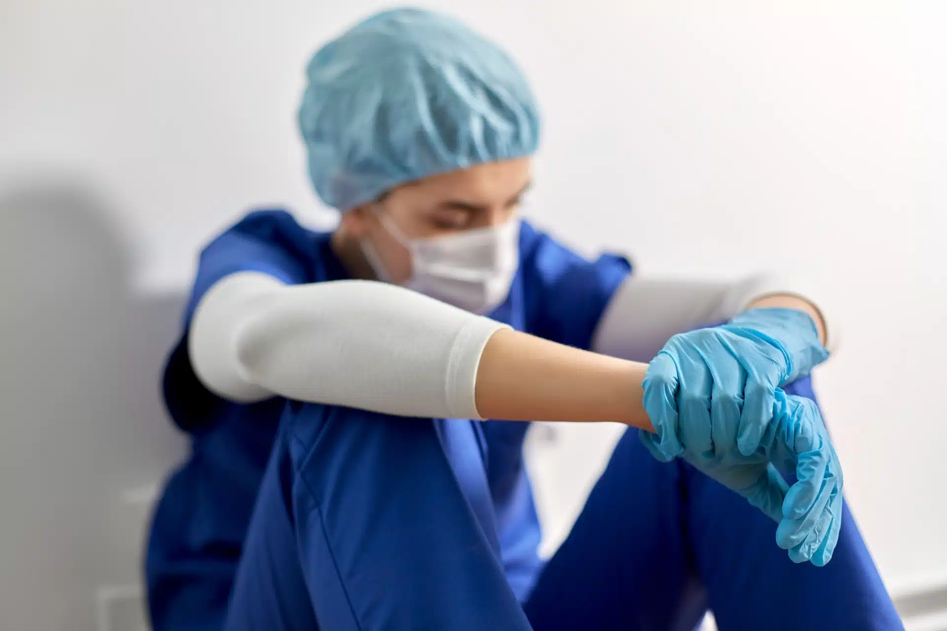 A nurse in blue scrubs and protective gear sits on the floor with her arms on her knees - chi franciscan health