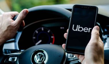 First-person view of an Uber driver sitting behind the wheel, looking at the Uber app on their phone - Uber drivers settlement