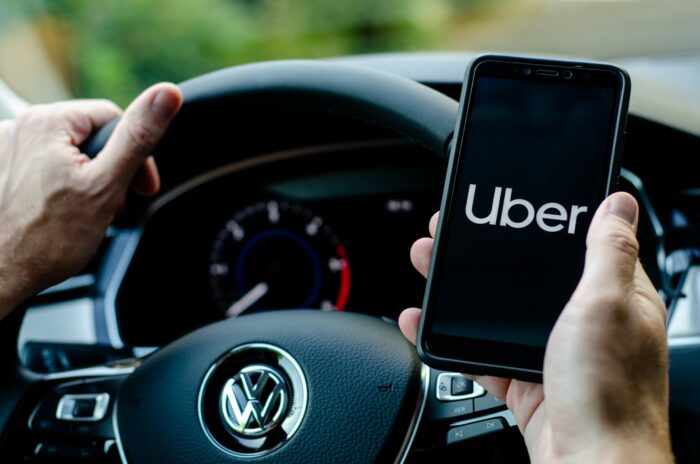 First-person view of an Uber driver sitting behind the wheel, looking at the Uber app on their phone - Uber drivers settlement - uber drivers data breach