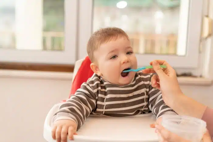 Young mother feeding her baby boy, whom sitting on the highchair, with a spoon. 
