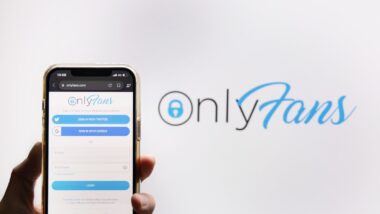 View of The Hand Holding a Mobilephone with Onlyfans Logo on the Screen Against Onlyfans Logo Background.