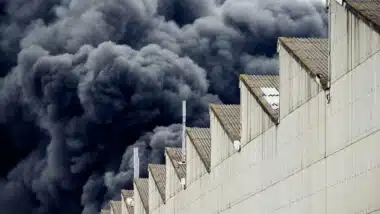 Black plumes of smoke from an accidental toxic industrial fire as seen from a behind a factory building.