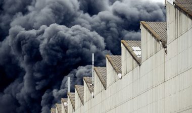 Black plumes of smoke from an accidental toxic industrial fire as seen from a behind a factory building.