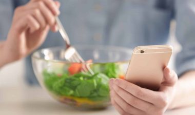 A woman uses a fork to eat salad from a glass bowl while holding a smartphone in her other hand - noom settlement