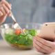A woman uses a fork to eat salad from a glass bowl while holding a smartphone in her other hand - noom settlement