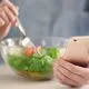 A woman uses a fork to eat salad from a glass bowl while holding a smartphone in her other hand - noom settlement
