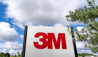 Close up of 3M sign with tree and blue sky in background.