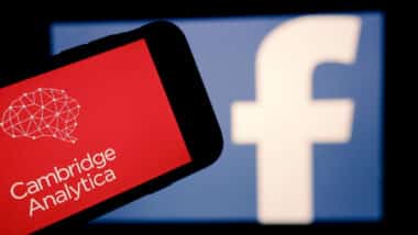 The logo of the strategic communication company Cambridge Analytica is seen on the screen of an iPhone in front of a computer screen showing Facebook logo.
